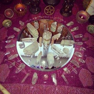 A top-down view of a psychic crystal grid arranged in a circular pattern on a reflective mirror base. Large clear quartz points and tumbled stones are meticulously placed over a purple patterned altar cloth, flanked by small bowls and lit votive candles for meditation and manifestation.